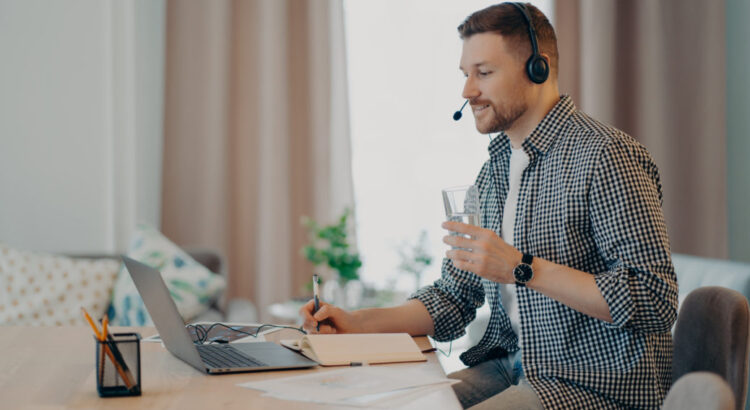 Male college student listens academic podcast sits at desk with laptop consults client by video makes notes drinks water from glass watches webinar dressed in checkered shirt poses at home office