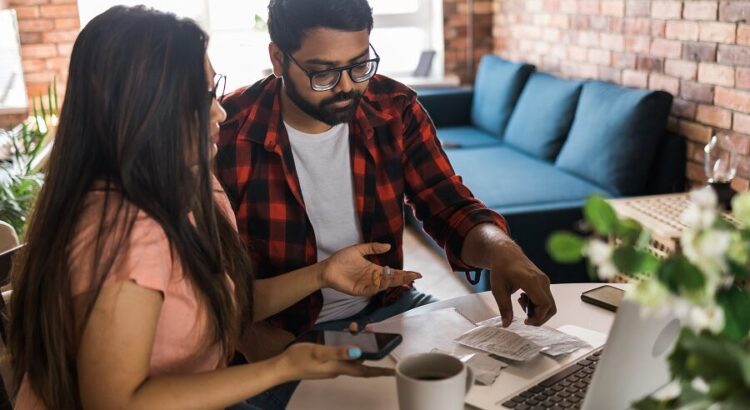 Young millennial indian husband and wife doing domestic paperwork, accounting job and reviewing paper bills, receipts at laptop computer, using online calculator and paying mortgage rent fees on internet - economic crisis concept